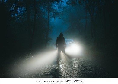 A sinister hooded figure standing in front of a car. On a spooky forest road on a foggy evening.