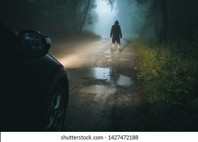 A Sinister Hooded Figure Standing In Front Of Car Headlights. On A Spooky Muddy Forest Road At Night.