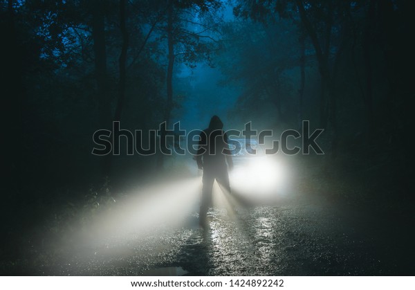 A sinister hooded figure\
standing in front of a car. On a spooky forest road on a foggy\
evening.