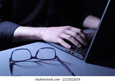 Sinister Looking Individual In Black Shirt Typing At Laptop With Glasses On Desk