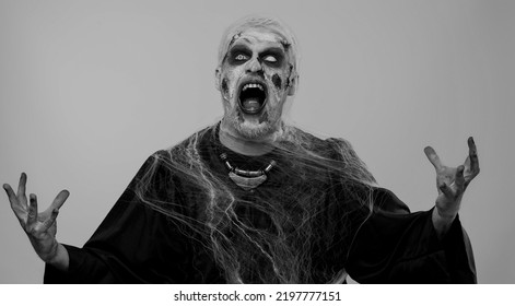 Sinister Man In Carnival Costume Of Halloween Crazy Zombie With Bloody Wounded Scars Face Screaming, Shouting, Trying To Scare On Gray. Horror Theme Of Cosplay Wounded Undead, Beast, Monster