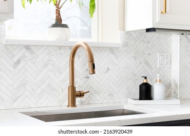 Sink detail shot in a luxury kitchen with herringbone backsplash tiles. white marble countertop, and gold faucet.