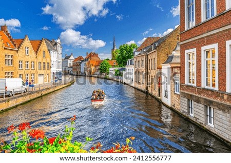 Sint annarei water canal with tourists motor boat, medieval houses on embankment, Brugge old town quarter in Bruges city historical centre, Church tower spire and flowers, Flemish Region, Belgium