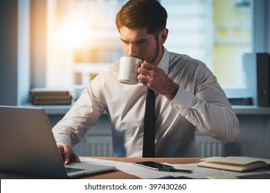 Sip Of Fresh Coffee At Work. Pensive Young Handsome Man Using His Laptop And Drinking Coffee While Sitting At His Working Place
