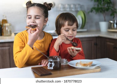 Sister And Brother Alone In The Kitchen Have Breakfast On Their Own Chocolate Paste, Smeared On The Toast Of Bread At The Table In The Kitchen. Children Have Fun Eating Chocolate Mousse