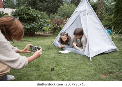 Sister and brother friendly play together in garden sitting in textile toy hut in summer day and their mother filming them using mobile phone - Powered by Shutterstock