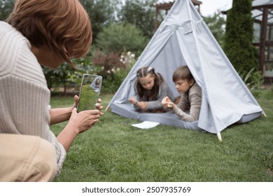 Sister and brother friendly play together in garden sitting in textile toy hut in summer day and their mother taking picture of them using phone - Powered by Shutterstock