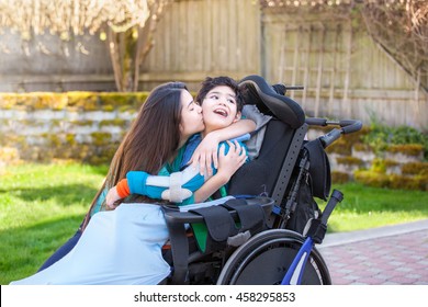 Sister kissing and hugging disabled little nine year old  brother in wheelchair outdoors