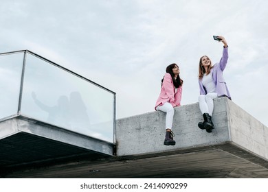 Sisters taking selfie through smart phone while sitting on rooftop in city against sky - Powered by Shutterstock