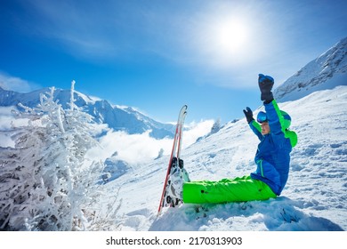 Site portrait of a boy with alpine ski sit in snow lifting hands, wear helmet and sport glasses on top of the mountain over peaks covered by clouds on background