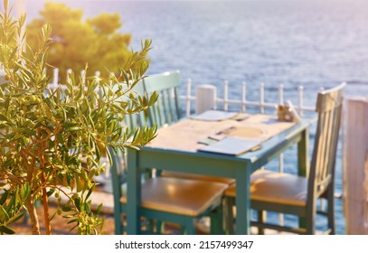 Sithonia, Chalkidiki, Greece. Traditional tavern cafe exterior with wooden table chairs. Olive tree branch focused in front with the view of the aegean sea in the background.