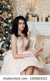 Sits On A Chair By The Fireplace And Holds A Glass Of Champagne. Beautiful Woman In Decorated Room. Christmas Celebration. Woman Decorating Christmas. Party Host Preparing For Holidays