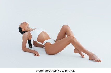 Sits on the floor. Woman with sportive slim body type in underwear that is in the studio.