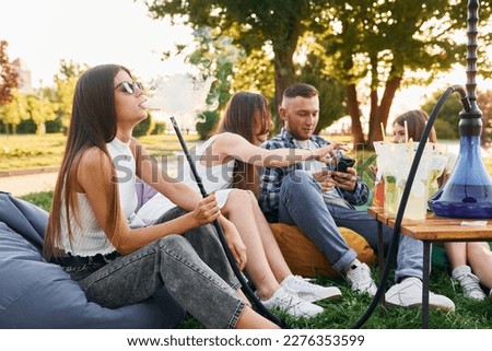 Sitting in the armchair bags and smoking hookah. Group of young people have a party in the park at summer daytime.
