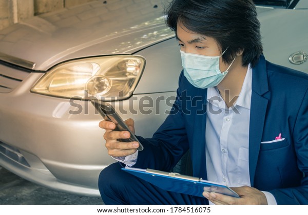 Sitting Asian Insurance Agent\
or Insurance Agency in Suit Wear Mask and See Smartphone and Hold\
Report Inspecting Car from Accident for Claim at Garage in Vintage\
Tone