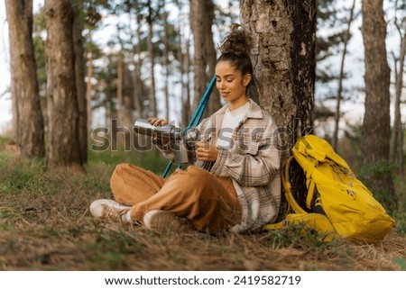 Sitting by the forest's border, the young woman with curly hair enjoys a peaceful coffee break while her trusty yellow backpack stands by.
