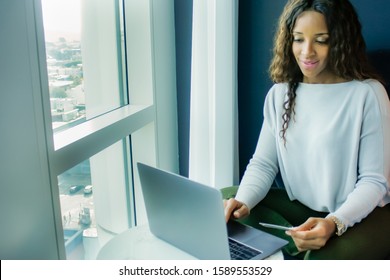 Sitting By A Large Window, A Beautiful African American Woman With Long Curly Hair And Credit Card In Hand, Makes A Purchase Online Using Her Laptop.