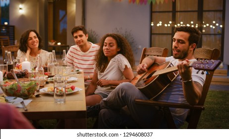 Sitting At The Dinner Table Handsome Young Man Plays The Guitar For A Friends. Group Of Young People Listening To Music At The Summer Evening Garden Party Celebration.