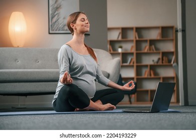 Sitting in lotus pose. With laptop. Beautiful pregnant woman is indoors at home. - Powered by Shutterstock