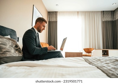 Sitting on the bed and working by using laptop. Businessman is indoors in the hotel room. - Powered by Shutterstock