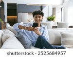 Sitting on couch, teenage boy smiling and using smartphone in modern home. Technology, mobile, communication, lifestyle, indoors, happiness