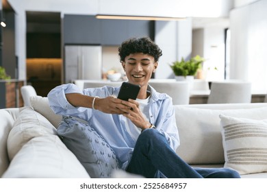 Sitting on couch, teenage boy smiling and using smartphone in modern home. Technology, mobile, communication, lifestyle, indoors, happiness - Powered by Shutterstock