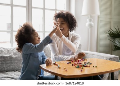 Sitting on sofa happy African little daughter gives high five to mother, family spend time together finish pastime activity, necklace jewelry from colorful wooden beads lie on table, hobby concept