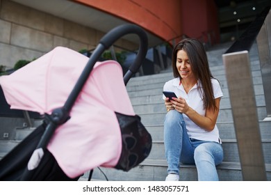 Sitting On The Stairs With Smartphone In Hands And Pink Pram Nearby. Pretty Young Woman Have Weekend In The City At Daytime.