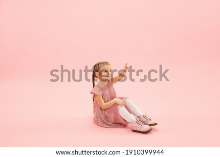 Sitting, pointing. Childhood and dream about big and famous future. Pretty longhair girl on coral pink studio background. Childhood, dreams, imagination, education, facial expression, emotions concept