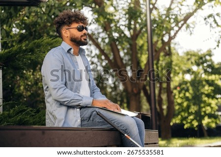 Sitting and reading using Braille. Blind man with white cane and sunglasses is in the city.