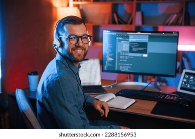 Sitting and smiling. Professional programmer is working indoors. Neon lighting.