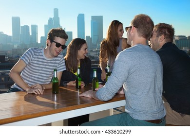 Six Adult Friends Talking And Drinking Beer At Rooftop Bar With Los Angeles Skyline, USA