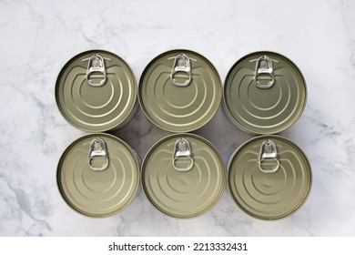 Six Cans Of Canned Food On A White Marble Kitchen Table. Canned Fish In A Tin Can. View From Above. Food Supplies, Donation Concept. Top View, Flat Lay