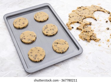 Six Chocolate Chip Cookies On A Metal Baking Tray, Raw Cookie Dough Scraps On Kitchen Table