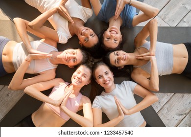 Six diverse funny girls lying together in circle on rubber mats wearing sportswear smiling show yoga greeting gesture, looking at camera, top above view. Namaste, symbol of salutation and valediction
