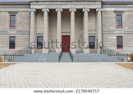 Six large round concrete columns at the top of marble steps with black iron rails to a legal building. The government building has a tall red door.  The view is from the lower corner of the steps.