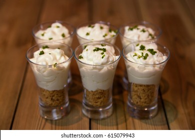 Six Mini Key Lime Pie Desserts In Small Glasses With Biscuits At The Base, Key Lime Pie Cream On Top And A Grated Lime Garnish On A Wooden Background With Shallow Depth Of Field And Space For Text.