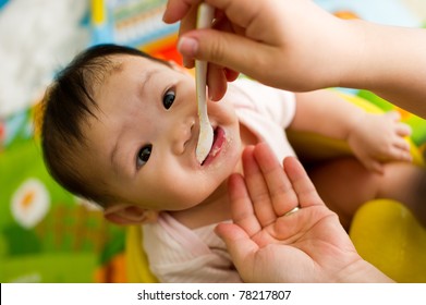 Six Month Old South East Asian Chinese Baby Girl Sitting In A Yellow Seat, Being Spoon Fed