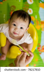 Six Month Old South East Asian Chinese Baby Girl Sitting In A Yellow Seat, Being Spoon Fed