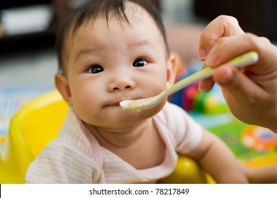 Six Month Old South East Asian Chinese Baby Girl Sitting In A Yellow Seat, Being Spoon Fed