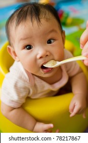 Six Month Old South East Asian Chinese Baby Girl Sitting In A Yellow Seat, Being Spoon Fed