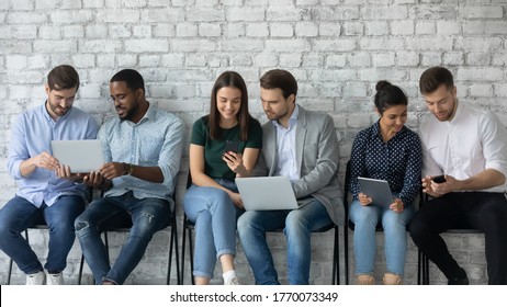 Six multi racial millennial girls and guys sit in line on chairs against grey wall using diverse devices, busy students discuss tasks, employees wait seminar in corridor having fun in internet concept - Powered by Shutterstock