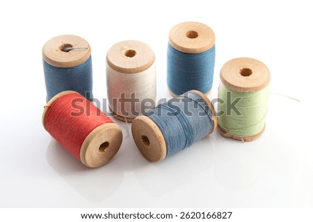 Six spools of vintage sewing thread in various colors sit on a white background. The spools have wooden tops and bottoms, showcasing a rustic charm. The threads are muted and appear well-worn