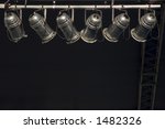 Six stage lights hanging in sunlight by dark wall and ladder