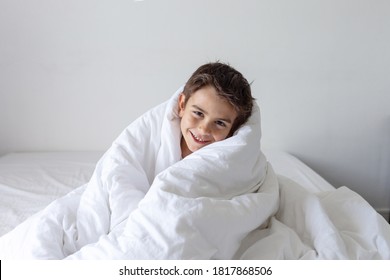 six year old boy wrapped himself in a white blanket while sitting on the bed and smiling