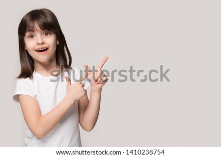 Six years old brown-haired little girl isolated on grey beige background, european appearance kid feel wondered look at camera pointing fingers aside at copy space freespace for your ad text concept