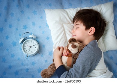 Six Years Old Child Sleeping In Bed On Pillow With Alarm Clock And A Teddy Bear