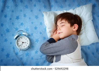 Six Years Old Child Sleeping In Bed On Pillow With Alarm Clock And A Teddy Bear