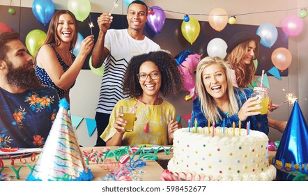 Six young adults celebrating a birthday party while holding drinks as they sit around a table with cake and party hats - Shutterstock ID 598452677