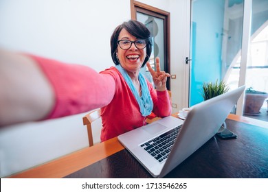 Sixty Year Old Female Teacher Wearing Headphones Taking A Selfihaving Online Class Via Video Chat On Laptop Computer. She Is Sitting On A Wooden Modern Desk At Home. Smiling And Enjoying Communication